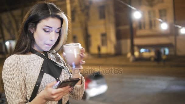 그녀의 스마트폰 및 겨울에 밖에 서 뜨거운 카 카오를 마시는 젊은 여자 문자 메시지 — 비디오