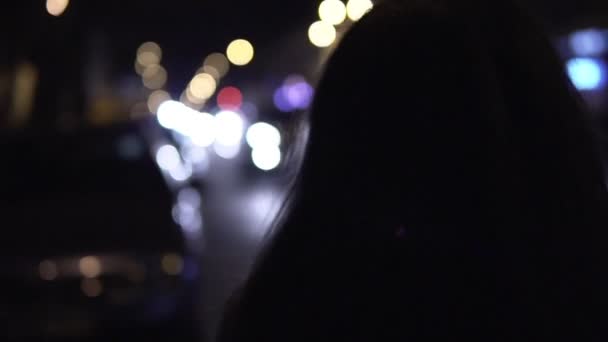 Jovem morena fica na rua à noite. Tráfego urbano em segundo plano — Vídeo de Stock