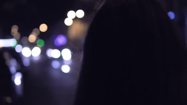 Молодая брюнетка стоит ночью на улице. Городское движение на заднем плане — стоковое видео