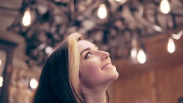 Young brunette woman looks up at the ceiling decorated with lamps and turns to camera — Stock Video