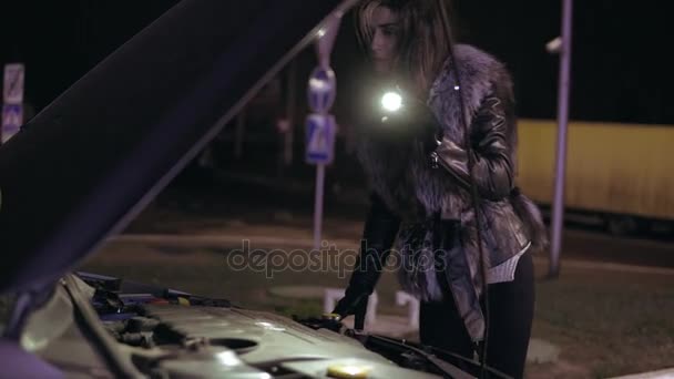 Young brunette woman with long hair is looking under the hood using her smartphone as a flashlight — Stock Video
