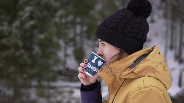 Jonge vrouw in zwarte bobbel hoed wandelingen in de buurt van de bomen in sneeuw drinken warme drank uit de papier-cup — Stockvideo