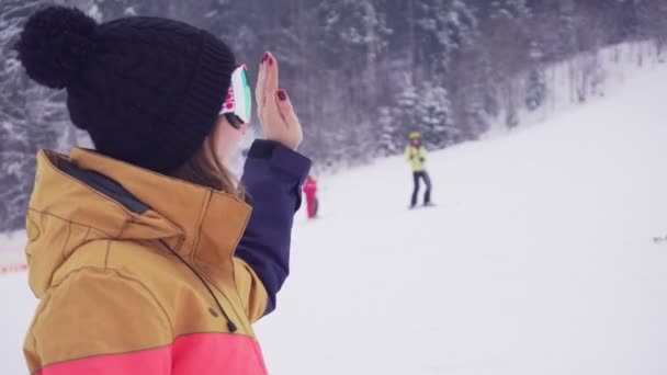 Молодая спортсменка в лыжных очках улыбается и машет рукой, называя кого-то — стоковое видео
