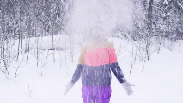 Young girl in bright winter clothes having fun outdoor in winter forest under snowflakes throwing up snow. — Stock Video