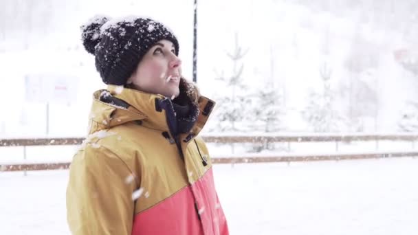 Молодая женщина в яркой зимней одежде и шляпе с шапкой стоит снаружи, пока идет сильный снег — стоковое видео
