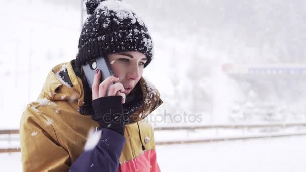 Νεαρή γυναίκα στο φωτεινό χειμωνιάτικα ρούχα μιλά στο τηλέφωνο έξω, ενώ χιονίζει σε μεγάλο βαθμό — Αρχείο Βίντεο