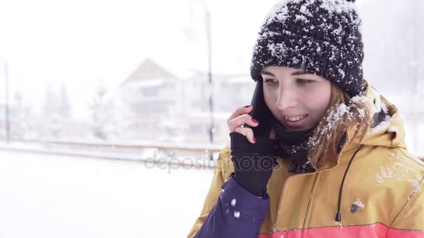 Mujer joven con ropa de invierno brillante habla por teléfono fuera mientras está nevando pesadamente — Vídeos de Stock