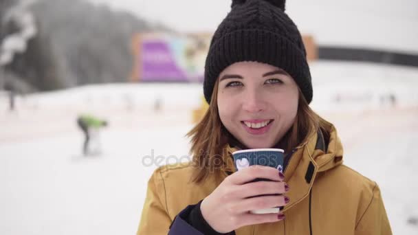 Chica con Taza de café para llevar sobre fondo de la estación de esquí. Hermosa mujer joven en sombrero de punto sonriendo. Retrato — Vídeos de Stock