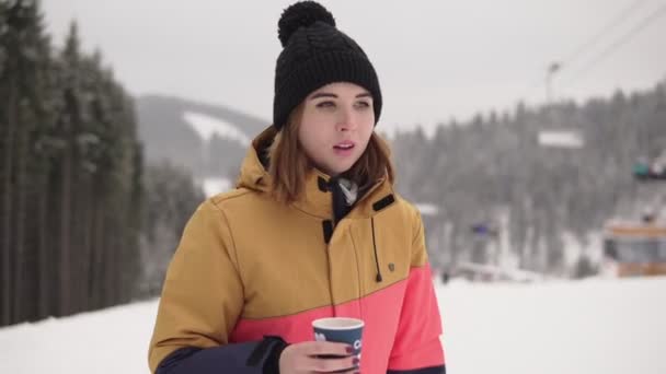 Fille avec tasse de café à emporter sur fond de station de ski marche sur la neige. Belle jeune femme en bonnet tricoté. Portrait — Video