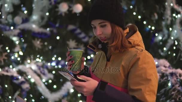 Jonge vrouw met kop hete thee met haar smartphone staande in de buurt van de kerstboom. — Stockvideo