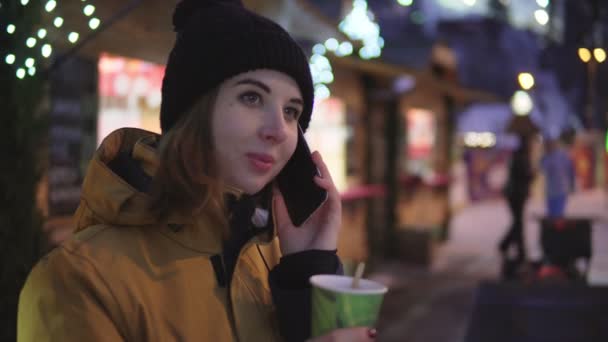 Schönes Mädchen, das eine Nummer wählt, mit einem Freund spricht und abends eine Papiertasse heißen Tee vor dem Christbaumhintergrund hält. — Stockvideo