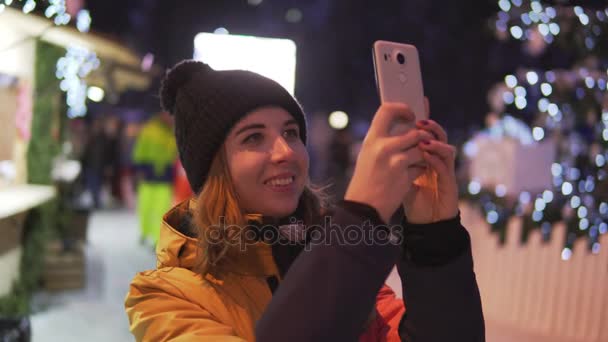 Молодая женщина в яркой зимней одежде фотографируется с мобильного телефона на Рождественском базаре — стоковое видео