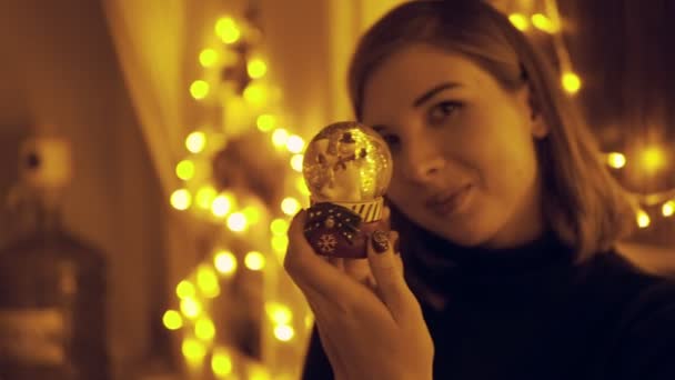 Portrait of a young woman with fairy lights in the house shaking the snow globe — Stock Video