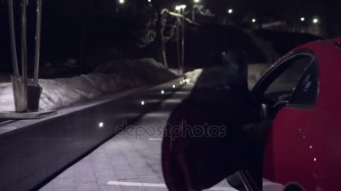 Genç adam gece Park, kırmızı arabadan alır ve kapıyı kapatır. Arkadan Görünüm.