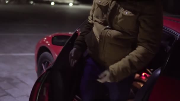 El joven se baja de un auto rojo por la noche en el estacionamiento. Vista trasera . — Vídeo de stock