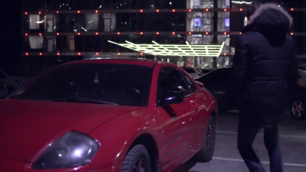 Hombre entrando en el coche deportivo rojo en el lado de los conductores. Luces de la ciudad en la noche en el fondo — Vídeo de stock