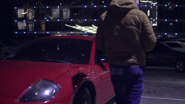 Мужчина садится в красную спортивную машину со стороны водителей. Городское освещение ночью на заднем плане — стоковое видео