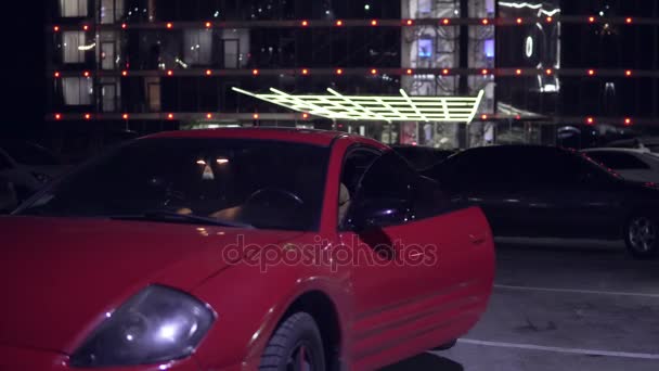 El joven sale de un coche rojo por la noche en el estacionamiento y cierra la puerta. Vista frontal. activar la señalización — Vídeo de stock