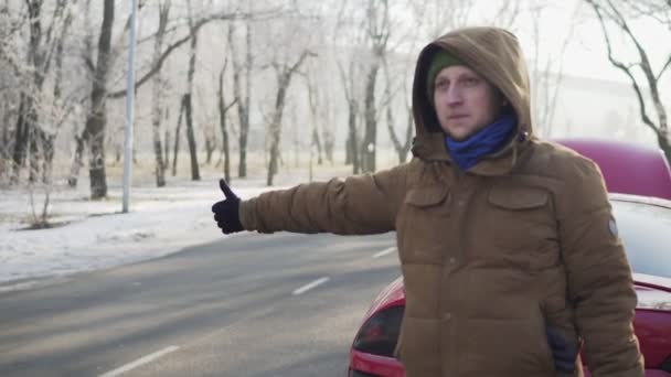 Attraktiv ung man lifta på en väg med sin uppdelade bil bakom honom — Stockvideo