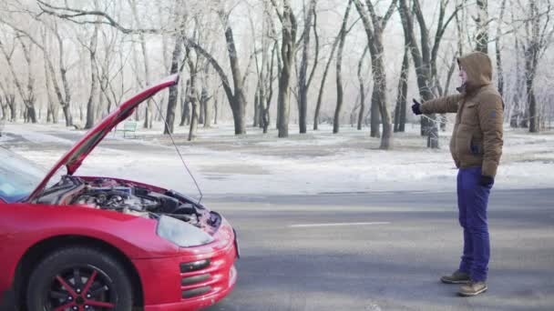 Attractive young man hitchhiking on a road with his broken down car close to him — Stock Video