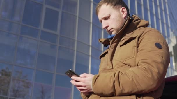 Jonge man zit door de auto, met zijn slimme telefoon, modern gebouw met grote ramen op de achtergrond — Stockvideo