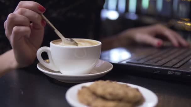 カプチーノと coockies のカップ。スプーンを使用してラップトップを背景に彼女のカプチーノを混乱させる女性. — ストック動画