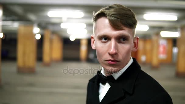 Vista de cerca del joven hombre rubio en un traje negro con una pajarita fumando un cigarrillo en el estacionamiento. Esperando a alguien . — Vídeo de stock