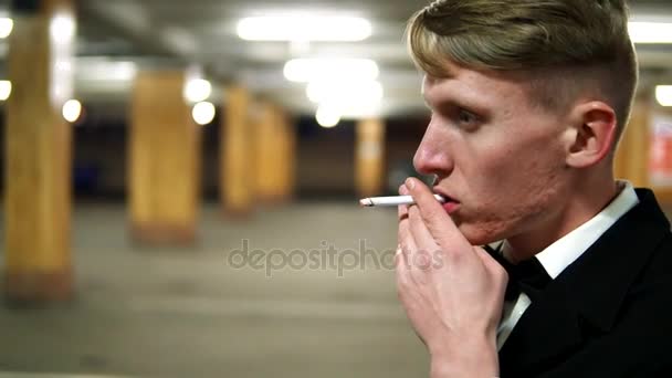 Vista de cerca del joven hombre rubio en un traje negro con una pajarita fumando un cigarrillo en el estacionamiento. Esperando a alguien . — Vídeo de stock