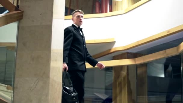 Молодой бизнесмен в костюме с бабочкой спускается по лестнице с большой черной кожаной сумкой. Слоумоция — стоковое видео