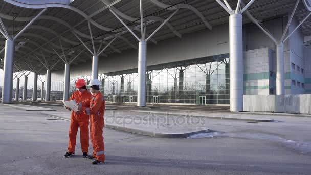 Οι εργαζόμενοι στις κατασκευές σε πορτοκαλί στολή και hardhats αναζητούν πάνω από σχέδια μαζί. Κτίριο στο παρασκήνιο — Αρχείο Βίντεο