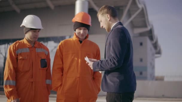 O gerente em um terno que dá envelopes com o dinheiro a funcionários no uniforme cor-de-laranja e capacetes. Acabamento bem sucedido do projeto. Movimento lento — Vídeo de Stock