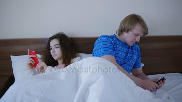 若い男と女の朝ベッドで横になっていると自分の携帯電話を見て — ストック動画