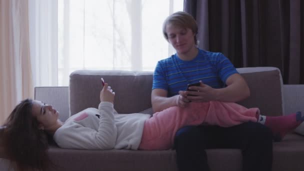 Young man and woman lying on the sofa and looking at their mobile phones, Man shows something on her phone to the woman — Stock Video