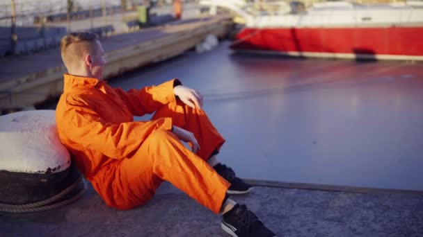 Mladý muž v oranžové uniformě sedí během své pauzy u moře v přístavu — Stock video