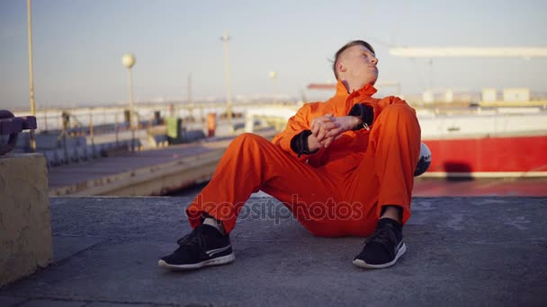 Joven en uniforme naranja sentado durante su descanso junto al mar en el puerto — Vídeos de Stock