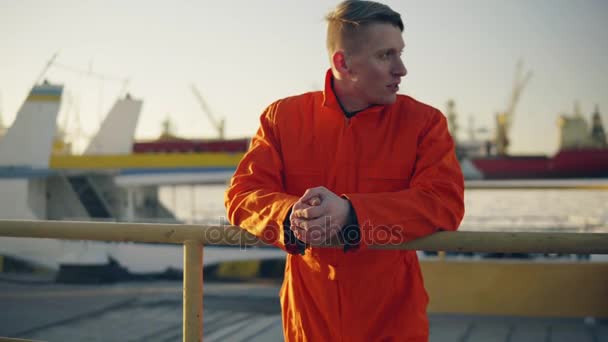 Young man in orange uniform standing by the fence and resting during his break by the sea in the harbor — Stock Video