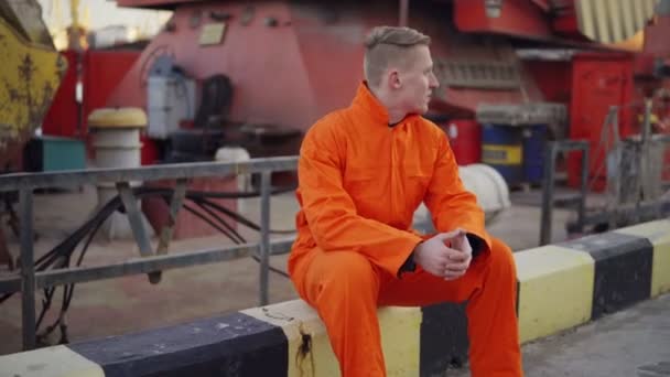 Joven en uniforme naranja sentado durante su descanso junto al mar en el puerto — Vídeo de stock