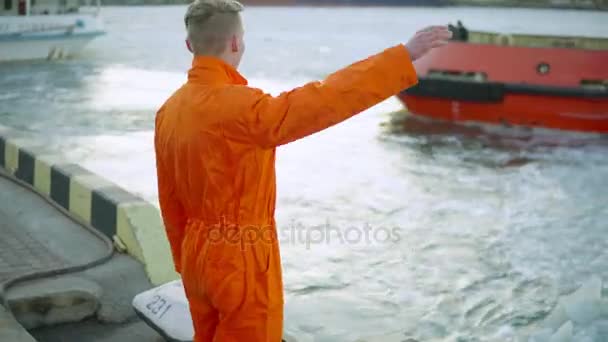 Young port worker in orange uniform waving his hand to the ship that is riding and going away — Stock Video