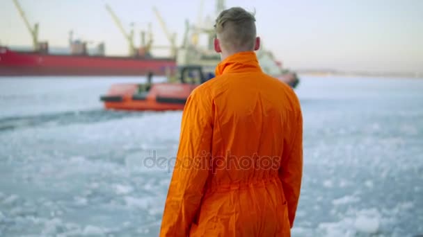 Trabajador portuario en uniforme naranja mirando al mar y caminando en el puerto en invierno. Mar helado — Vídeos de Stock