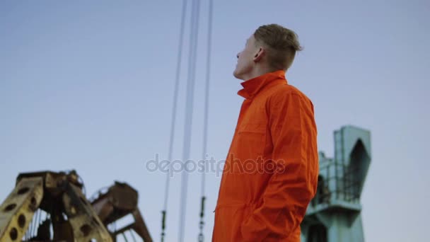 Красивый молодой работник контейнерного склада в оранжевой форме стоит у корабля в гавани и смотрит вверх — стоковое видео