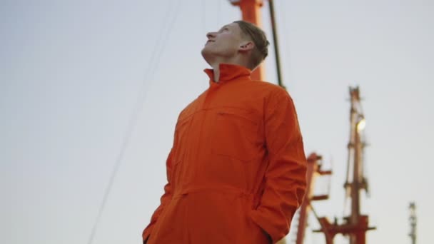 Красивий молодий працівник контейнерного складу в помаранчевій формі стоїть біля корабля в гавані і дивиться вгору — стокове відео