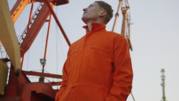 オレンジ色の制服と見上げると港の船のそばのハンサムな若いコンテナー倉庫労働者。背景に大きなクレーン. — ストック動画