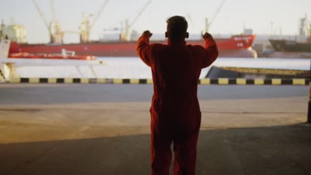 Silueta de un trabajador de uniforme naranja caminando por el almacén del puerto junto al mar durante su descanso y levantando las manos. Joven feliz . — Vídeo de stock
