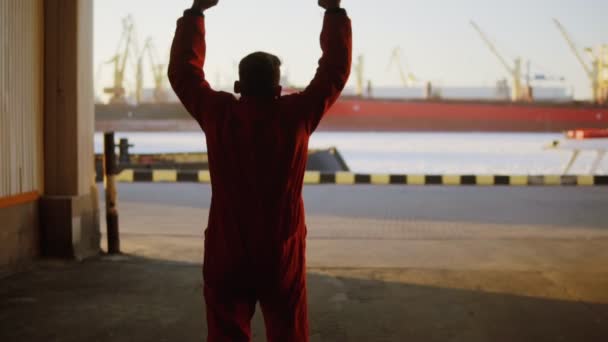 Silhouette of a worker in orange uniform walking through the harbour storage by the sea during his break and raising his hands up. Happy young man. — Stock Video