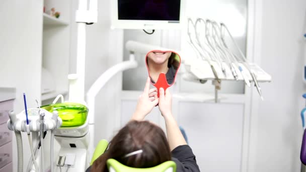 Joven mujer hermosa en la silla dental. Después del procedimiento se mira en el espejo. El concepto de una sonrisa saludable . — Vídeo de stock