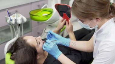 Prosedür sonuçlarını kontrol etmek için bir ayna tutan diş hekimi, genç bayan hasta. Genç kadın dişçi eldiven ve maske araçlarıyla yaklaşıyor.