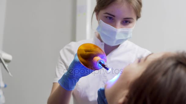 Jovem dentista feminina em máscara e luvas usando equipamento de luz UV dental para endurecimento de polímeros — Vídeo de Stock