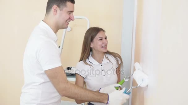 Cheerful young couple in white shirts decorate their new apartment. Young husband and wife are painting the wall using paint rollers. Beautiful couple making repairs in their new flat. — Stock Video