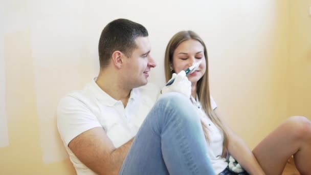 Young couple taking a break from renovating and laughing, sitting on the floor. Man is painting his wifes nose — Stock Video