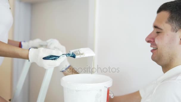Jovem alegre em camisa branca pintando a parede na cor branca usando um pincel e de pé na escada. Jovem marido está ajudando — Vídeo de Stock